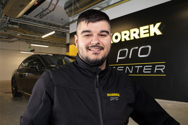 Hymyilevä Korrek Pro Center -asiantuntija seisoo autohallissa taustallaan kiiltävä auto ja brändiseinä.